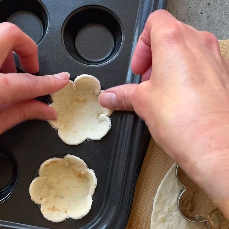 Hands pressing flower-shaped tortilla pieces into a muffin tin for a delightful avocado appetizer, with a wooden board and metal cutter visible nearby.