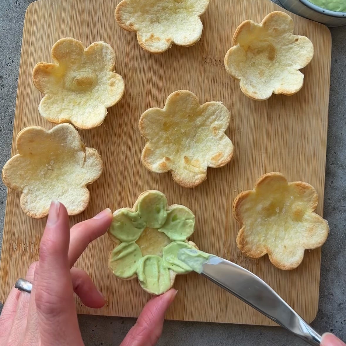 Seven flower-shaped tortilla cups on a wooden board. A hand spreads creamy avocado filling inside one cup with a butter knife.