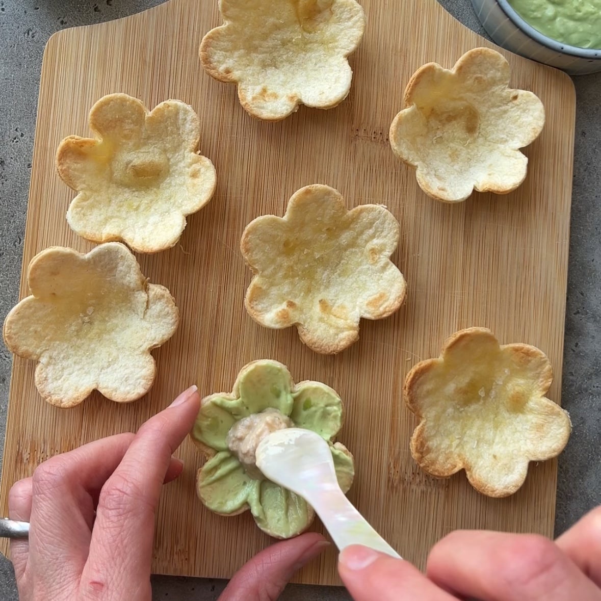 Seven flower-shaped tortilla flowers on a wooden board, with a hand spreading tuna salad into one shell using a spoon.