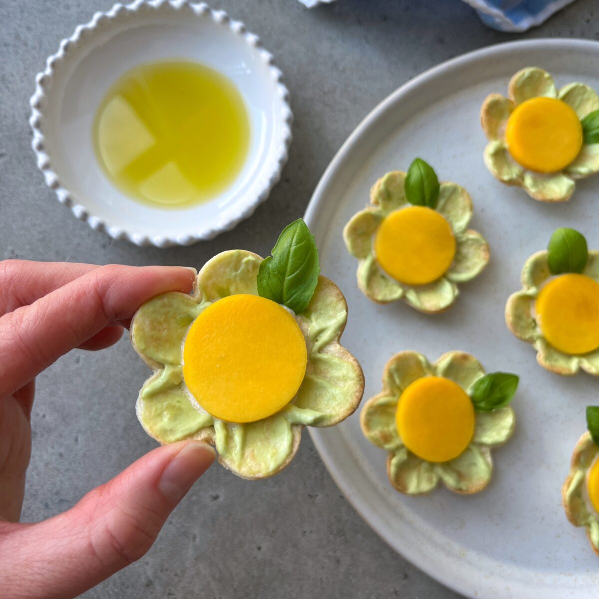 A hand holds a flower-shaped avocado appetizer with a yellow mango center and basil leaf. More appetizers rest on a white plate, with a small bowl of olive oil nearby.