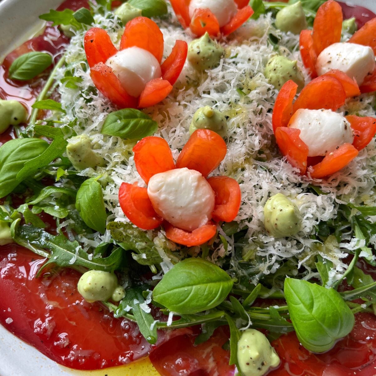 A bresaola salad with arugula, sliced tomatoes, basil, grated cheese, and decorative tomato flowers filled with mozzarella, topped with green herb dressing.