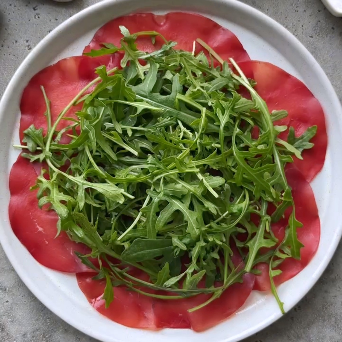 A platter is topped with beef carpaccio slices in a circular pattern, and fresh, vibrant arugula.