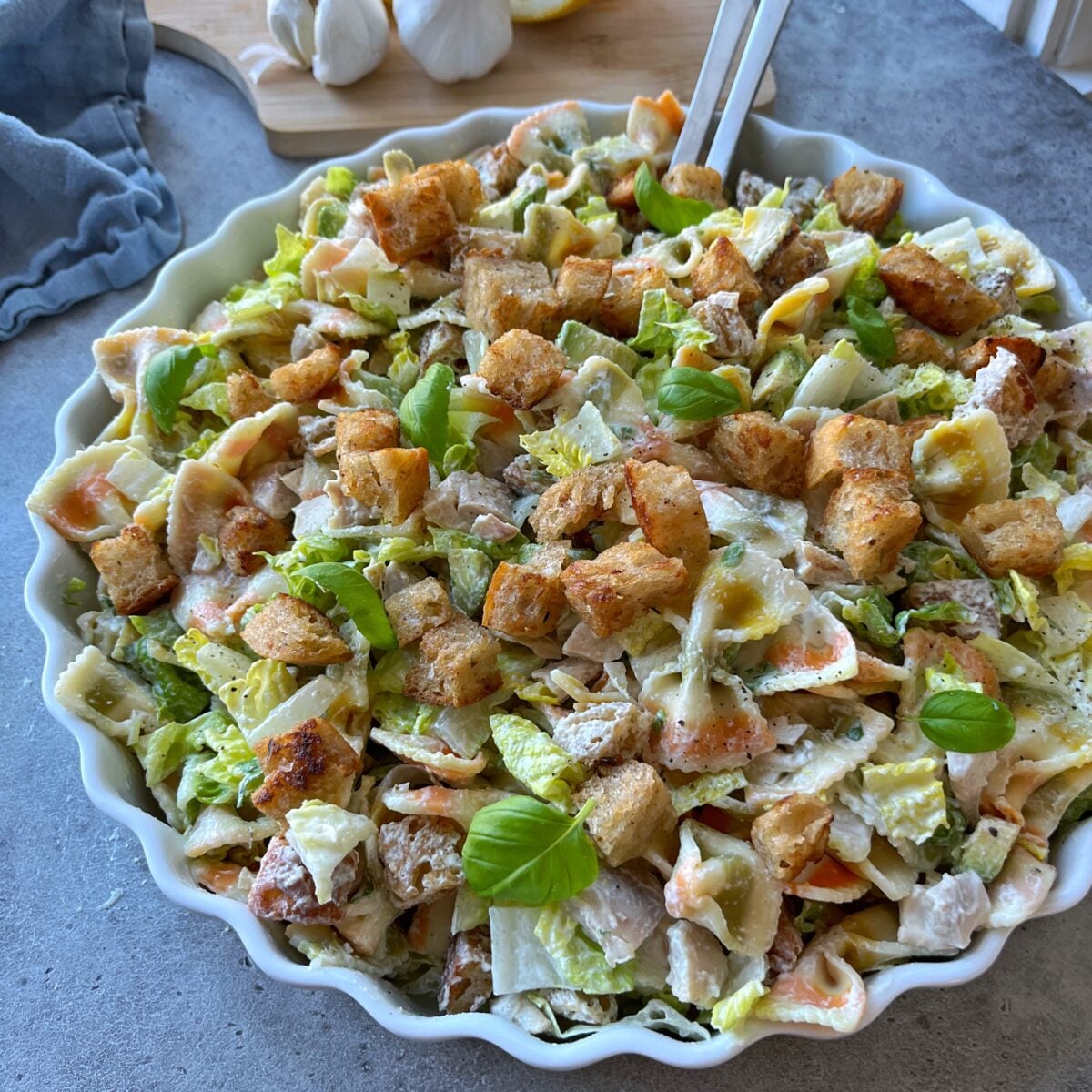 A bowl of Chicken Caesar Pasta Salad with lettuce, croutons, basil leaves, and creamy dressing on a gray surface.
