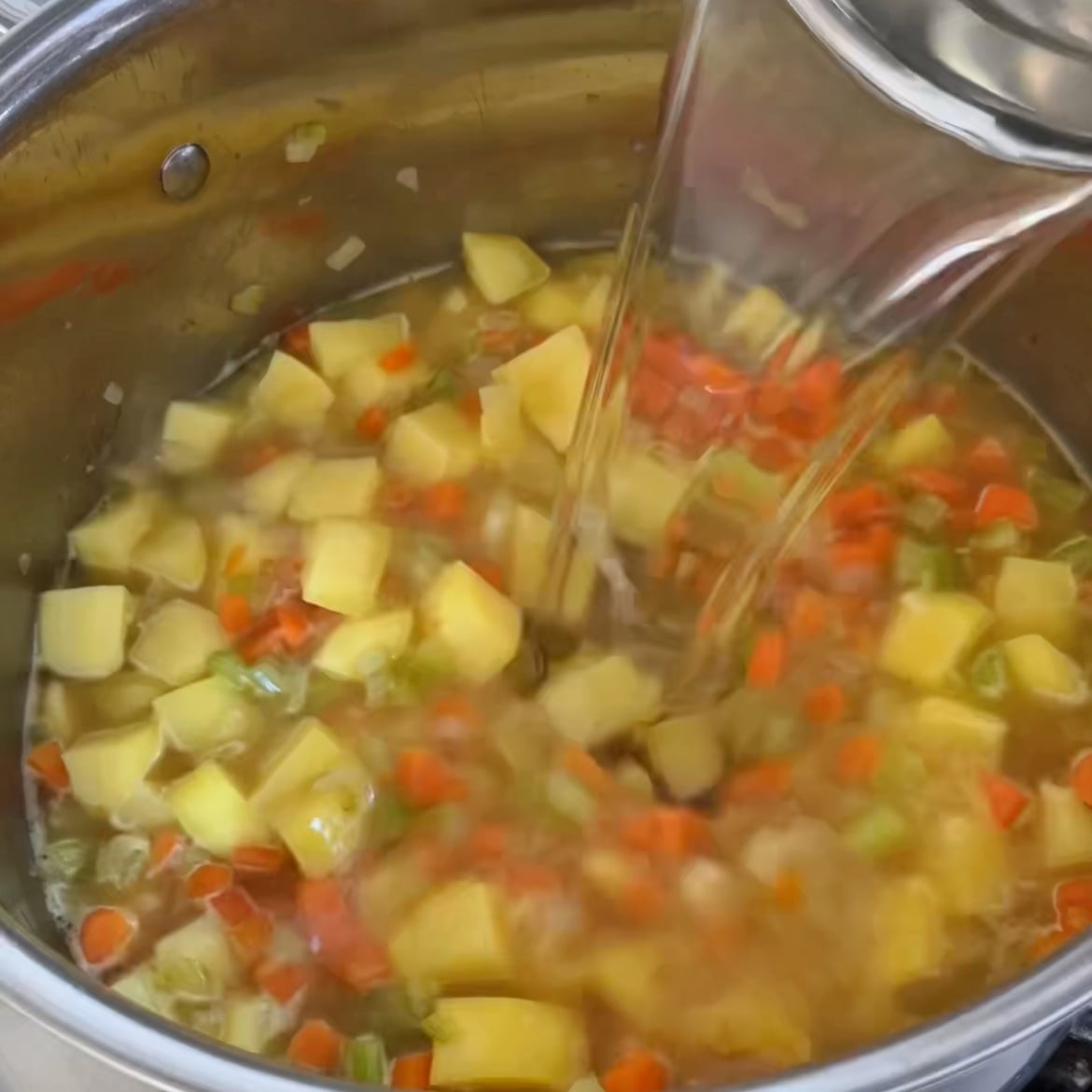 Water is being poured into a pot containing chopped potatoes, carrots, celery, and onions.