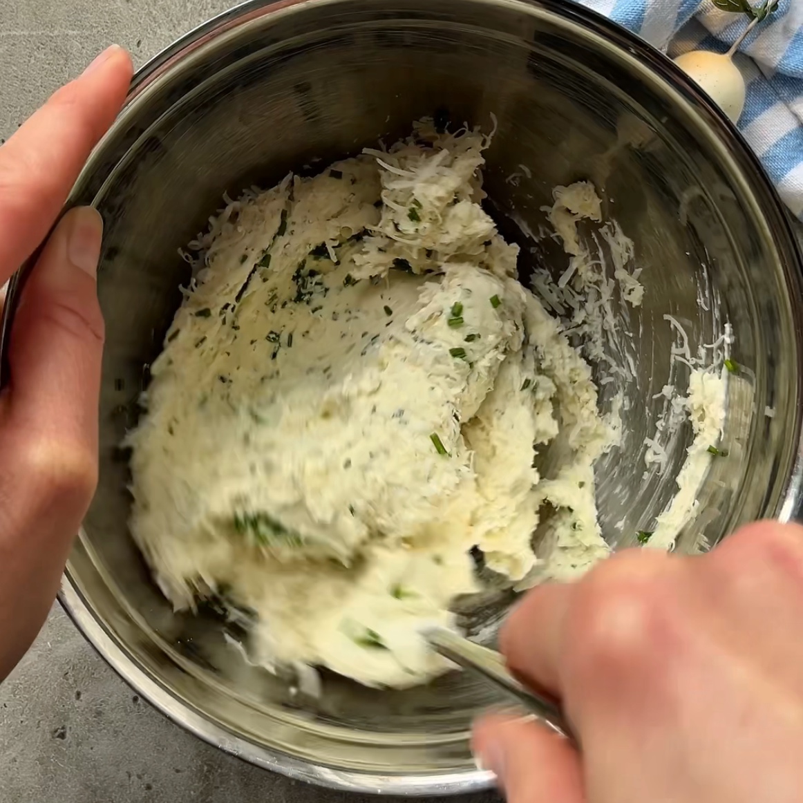 A person mixing an herbed Easter Cheese Ball mixture in a stainless steel bowl with a fork on a gray countertop.