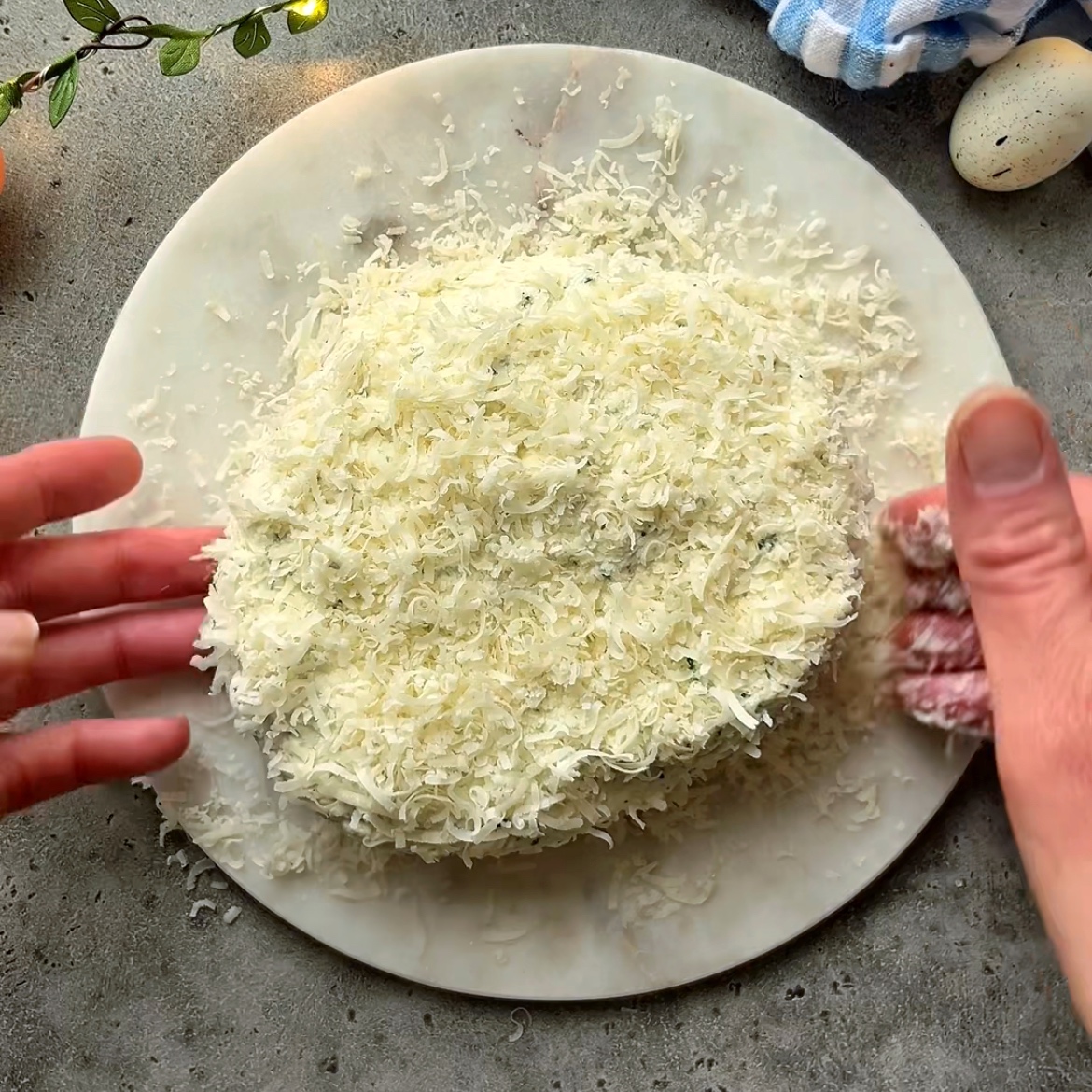 A person coats an Easter Cheese Ball with grated cheese on a white marble plate, surrounded by egg fairy lights and a striped cloth on a gray surface.