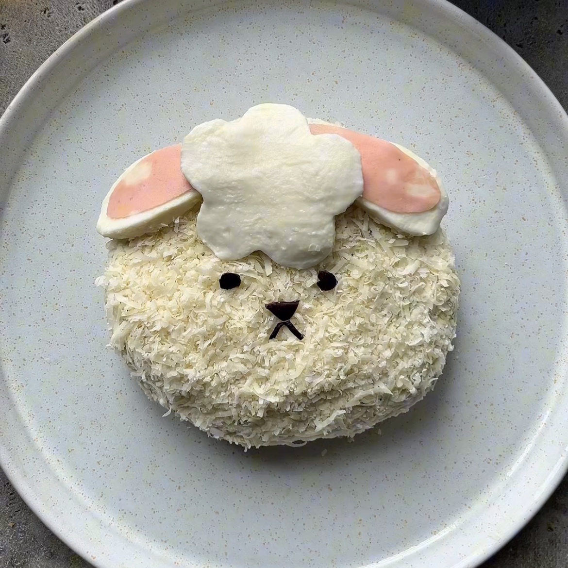 A round Easter Cheese Ball dessert decorated to look like a white lamb face with pink ears, olive eyes, and a small mouth, placed on a plain white plate.