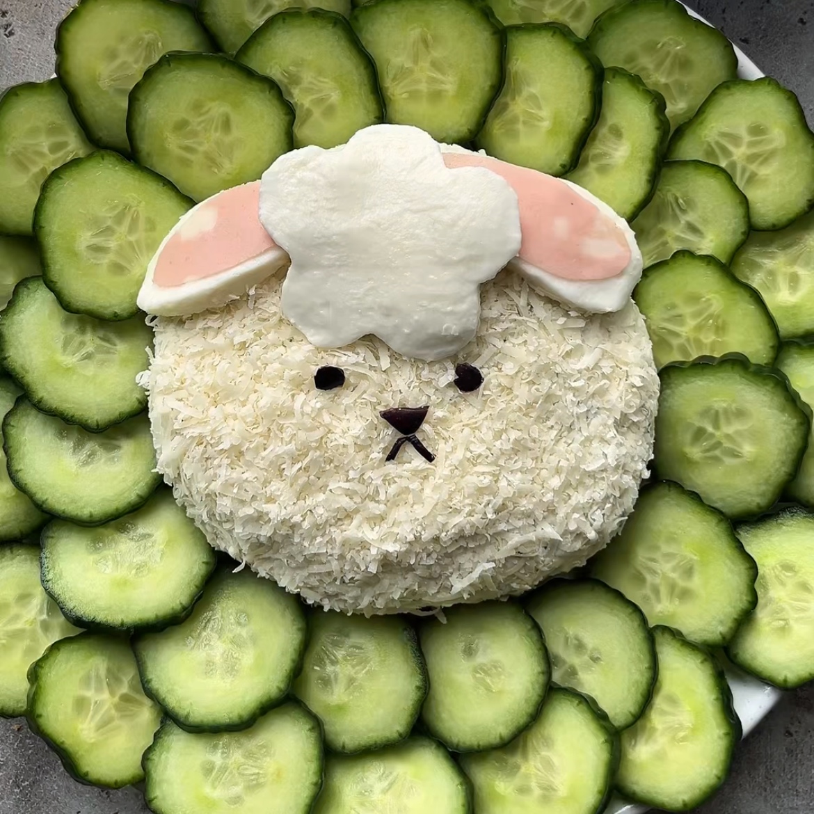 A festive Easter Cheese Ball, shaped like a lamb face, sits at the center of a plate surrounded by neatly overlapping cucumber slices.