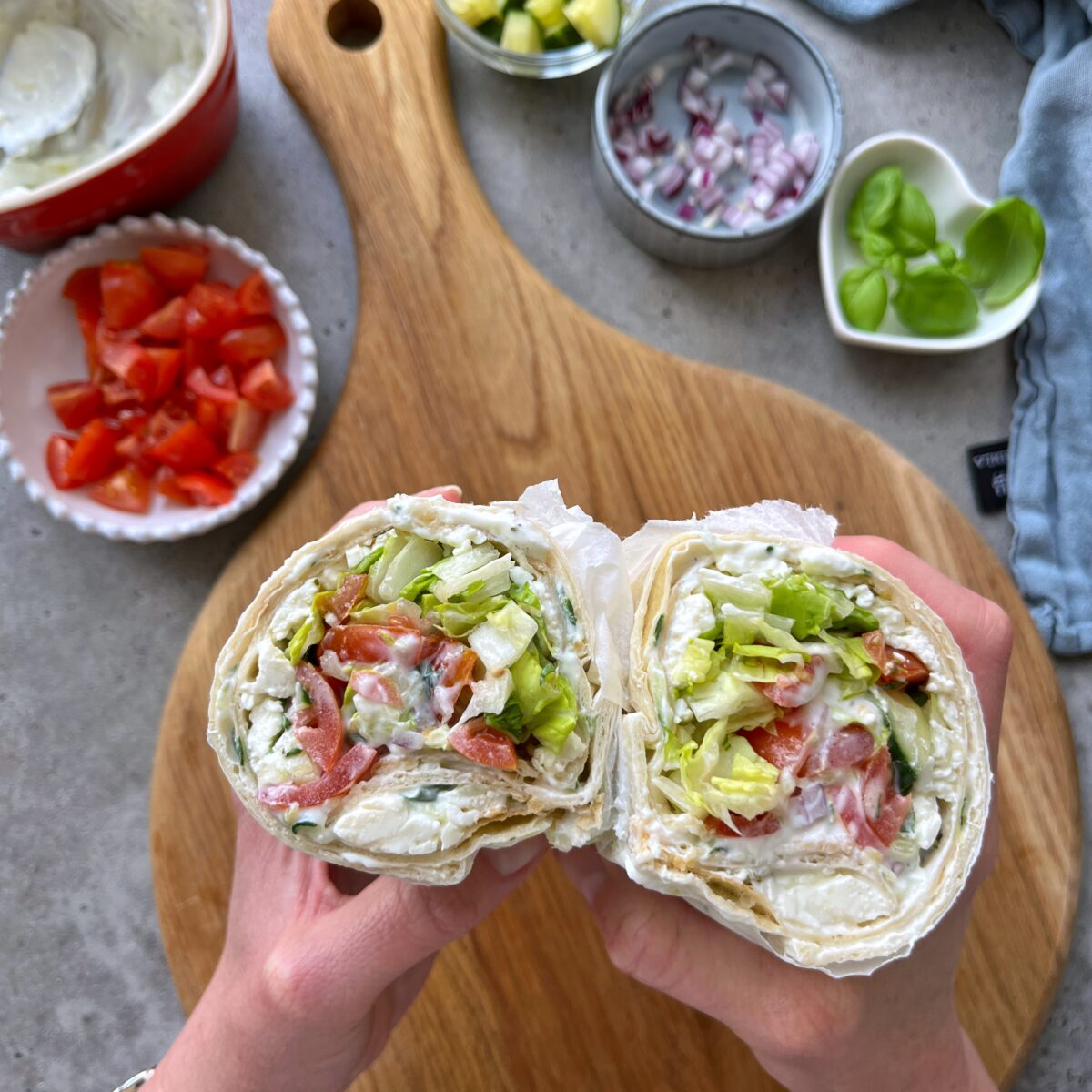 Two hands hold a sliced Greek Salad Wrap filled with lettuce, tomato, creamy spread, and other vegetables above a wooden cutting board with toppings in small bowls.