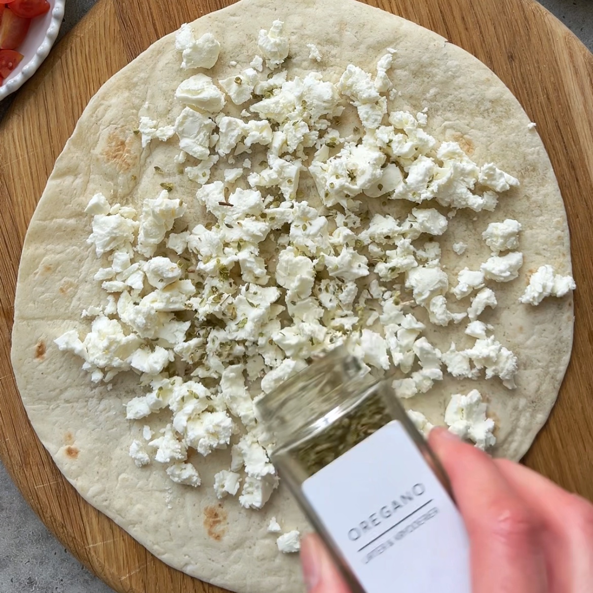 A hand sprinkles dried oregano from a jar onto a tortilla topped with crumbled feta cheese and placed on a wooden board.