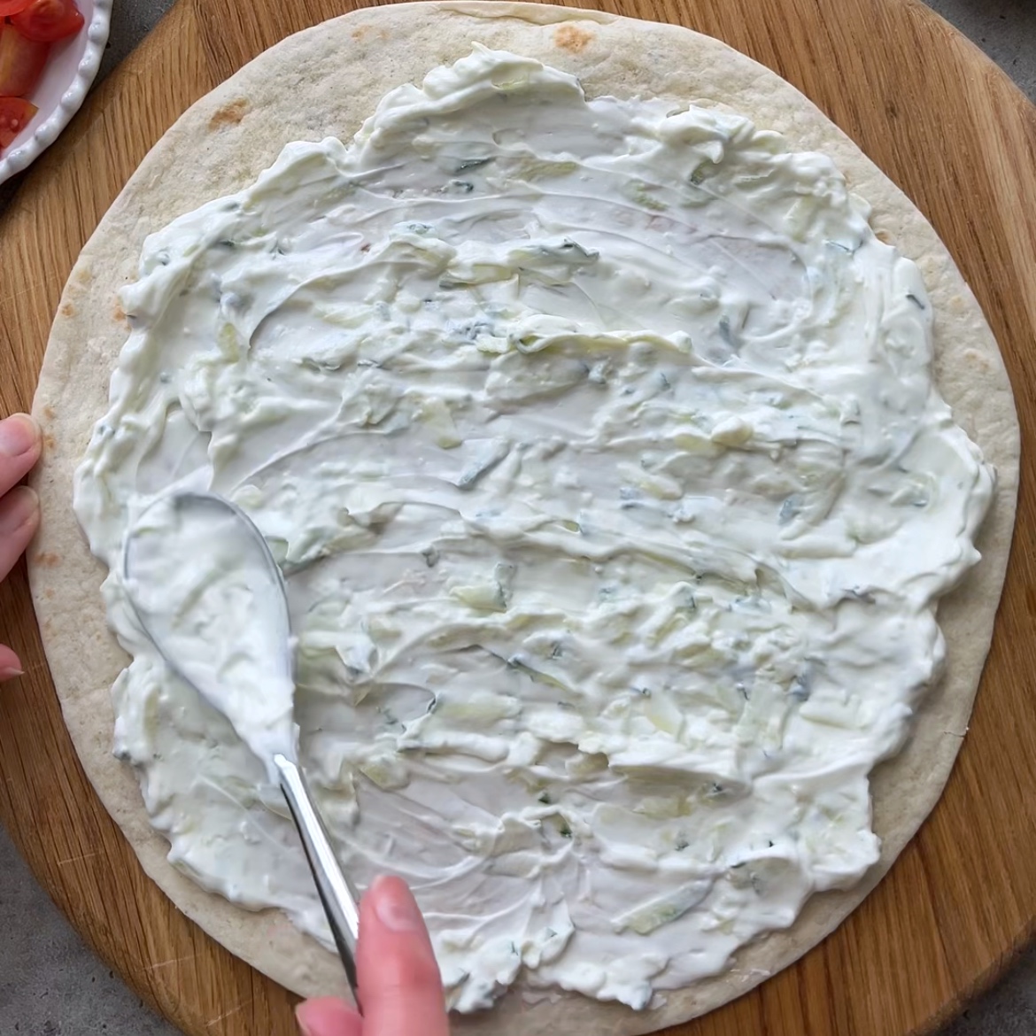 A hand spreads a creamy tzatziki over a round tortilla.