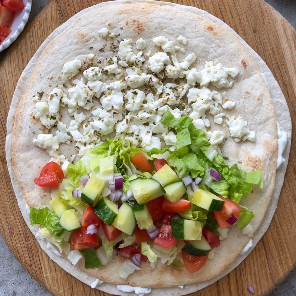 A tortilla on a wooden board topped with crumbled cheese on one half and chopped lettuce, tomato, cucumber, and red onion on the other half.