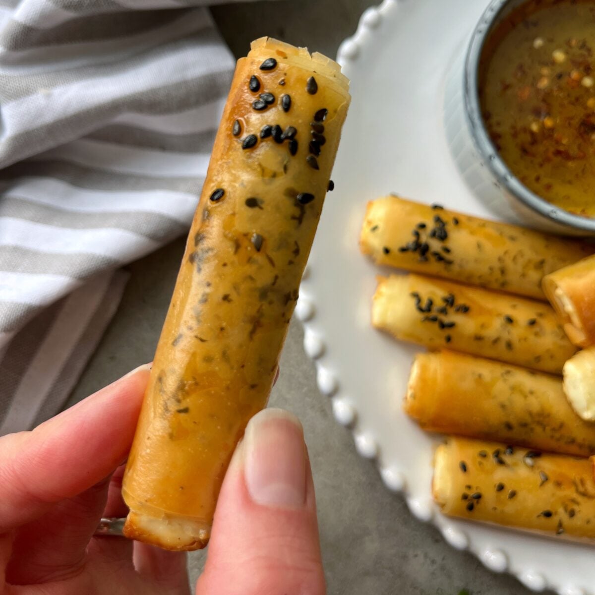 A hand holds a crispy, sesame-topped phyllo fry roll near a plate of similar rolls and a bowl of dipping sauce.