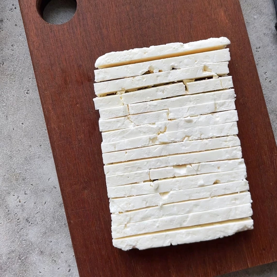 A block of white feta is evenly sliced and placed on a rectangular wooden cutting board.