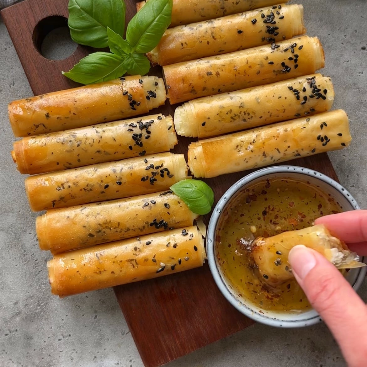 Crispy Phyllo Feta Fries topped with black sesame seeds are arranged on a wooden board with basil leaves, next to a bowl of hot honey.
