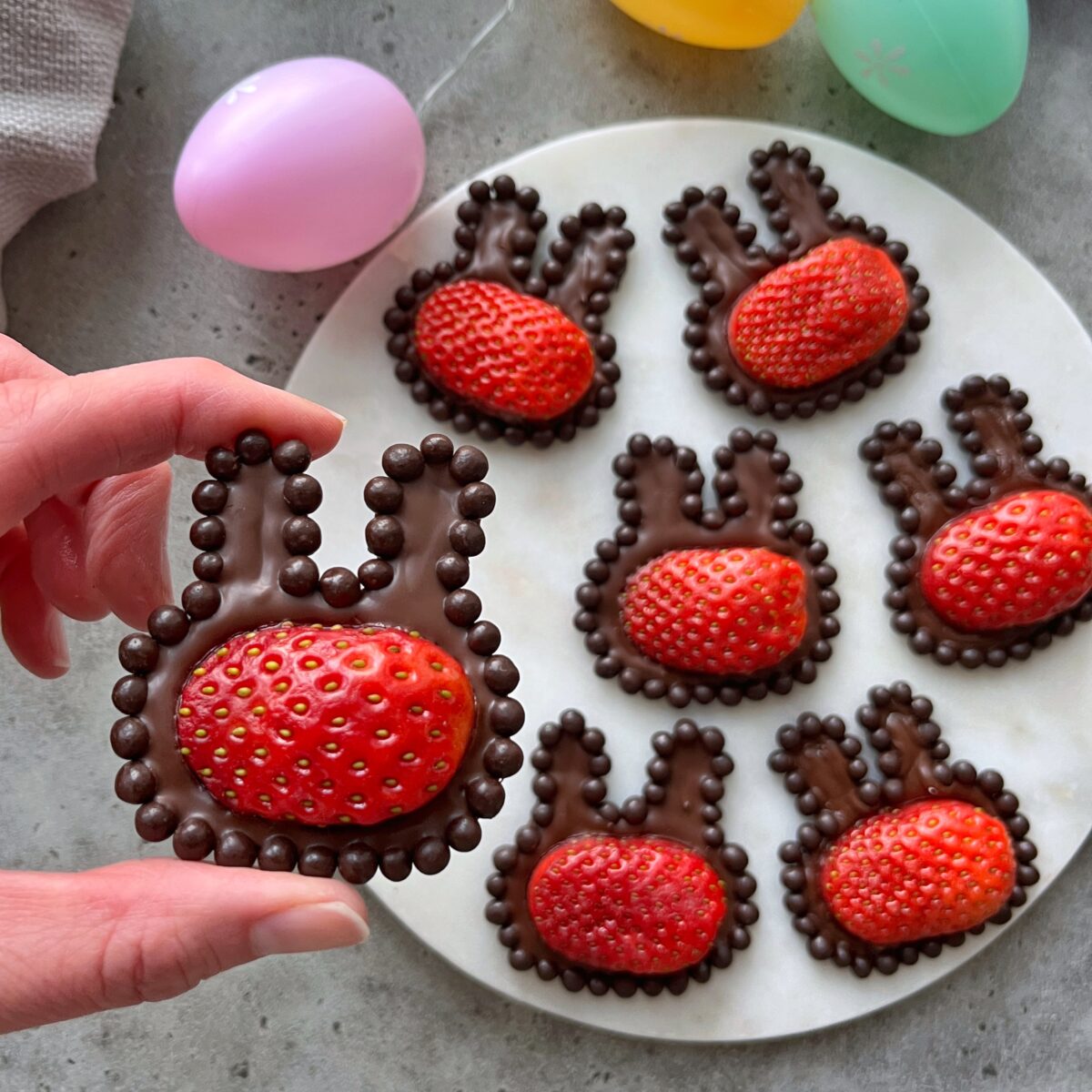A hand holds a chocolate Strawberry Bunnies treat topped with a fresh strawberry, while similar bunny-shaped delights are arranged on a round white plate.