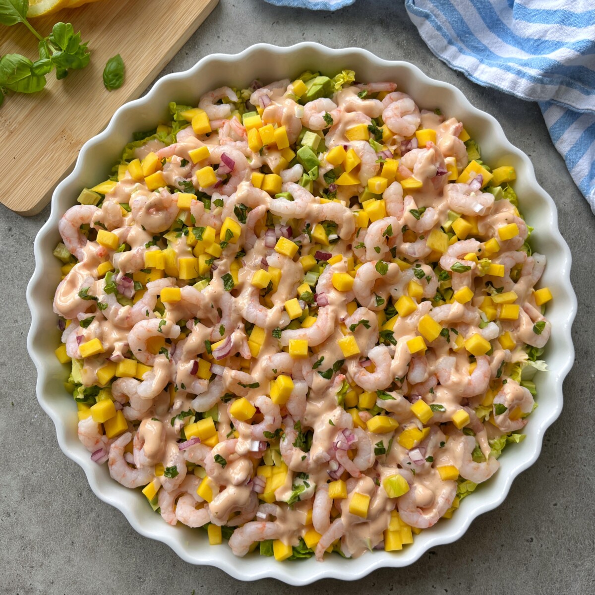 A round dish filled with a shrimp cocktail salad topped with diced mango, avocado, red onion, and a creamy pink dressing, garnished with chopped herbs. A blue-striped towel and cutting board with basil are nearby.