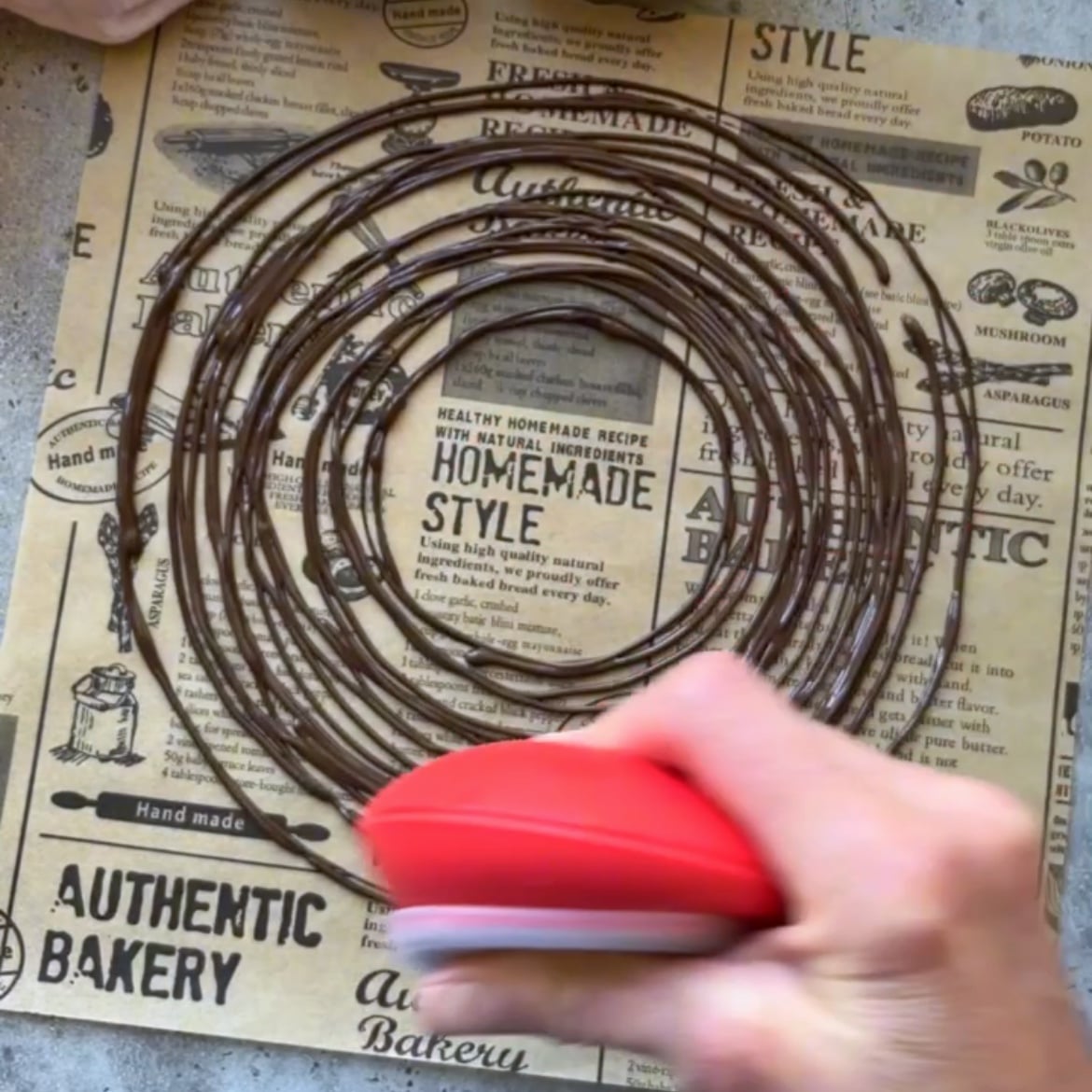A hand holding a red piping tool spreads chocolate on parchment paper.