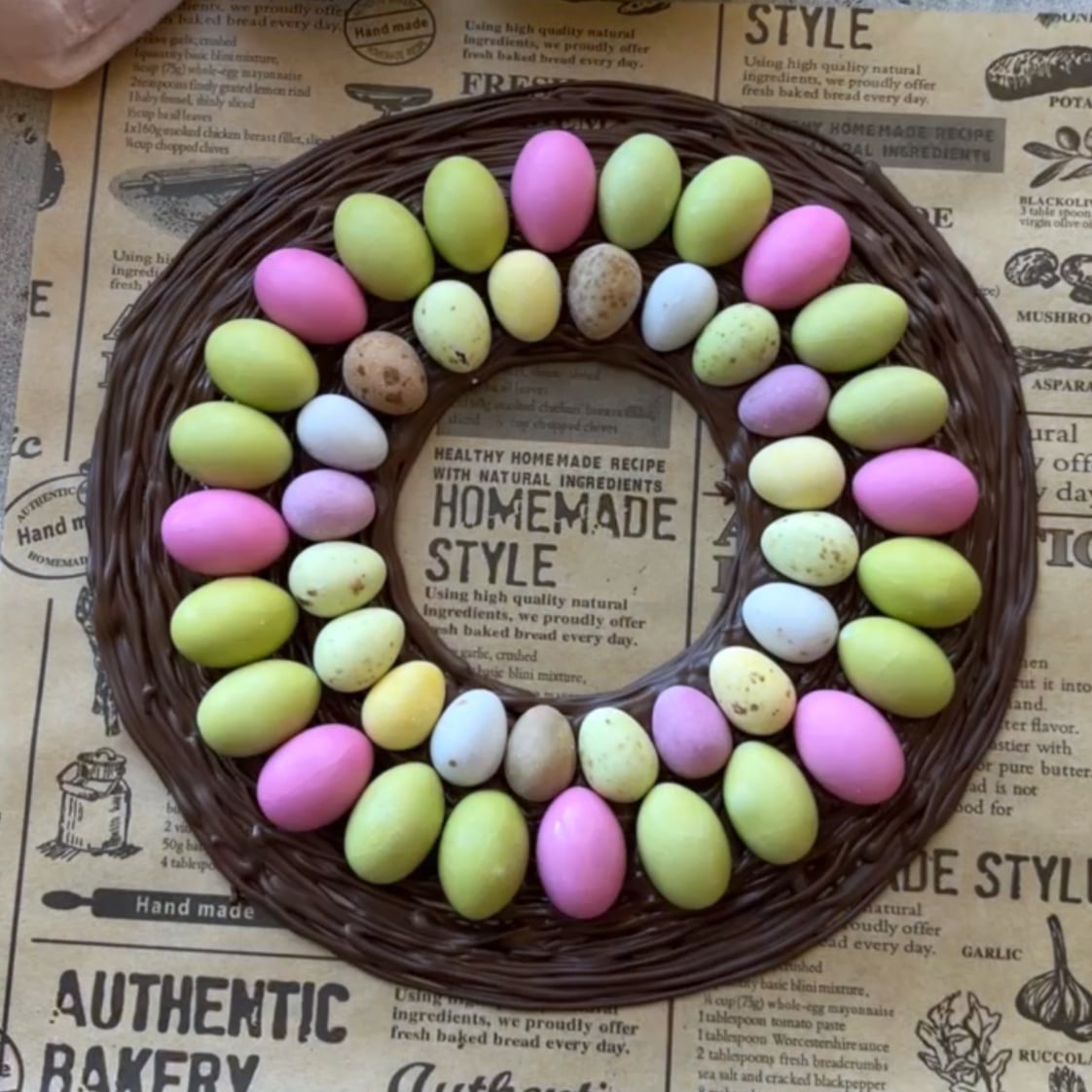 A circular wreath made of chocolate is decorated with pastel-colored and speckled egg-shaped candies, arranged in two rings on a printed paper background.