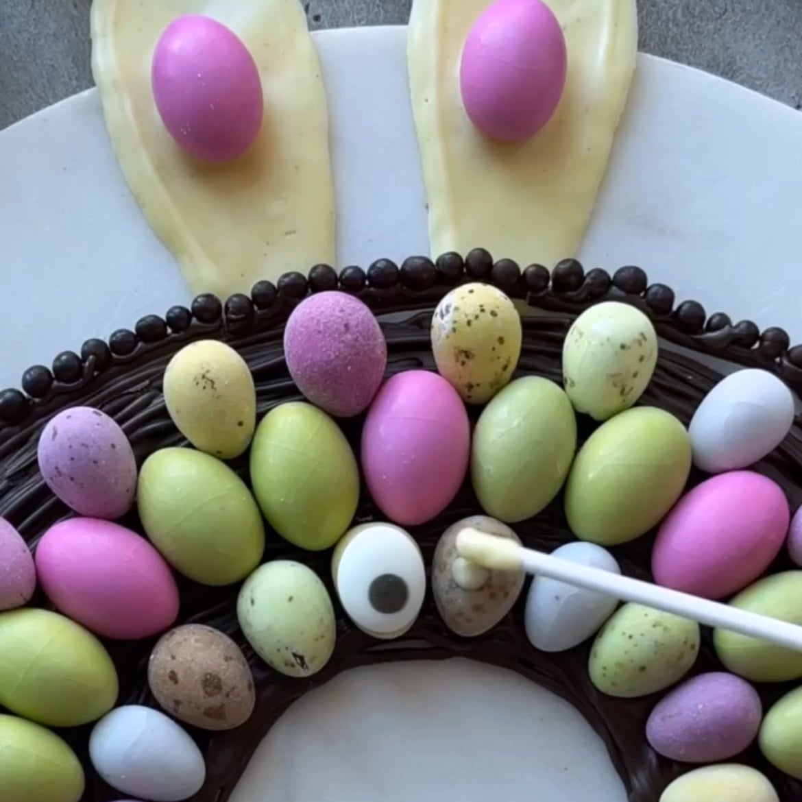 A decorative Easter Bark wreath made of chocolate and pastel candy eggs, topped with two white chocolate bunny ears. A hand is adding a candy eye detail with a lolly stick.