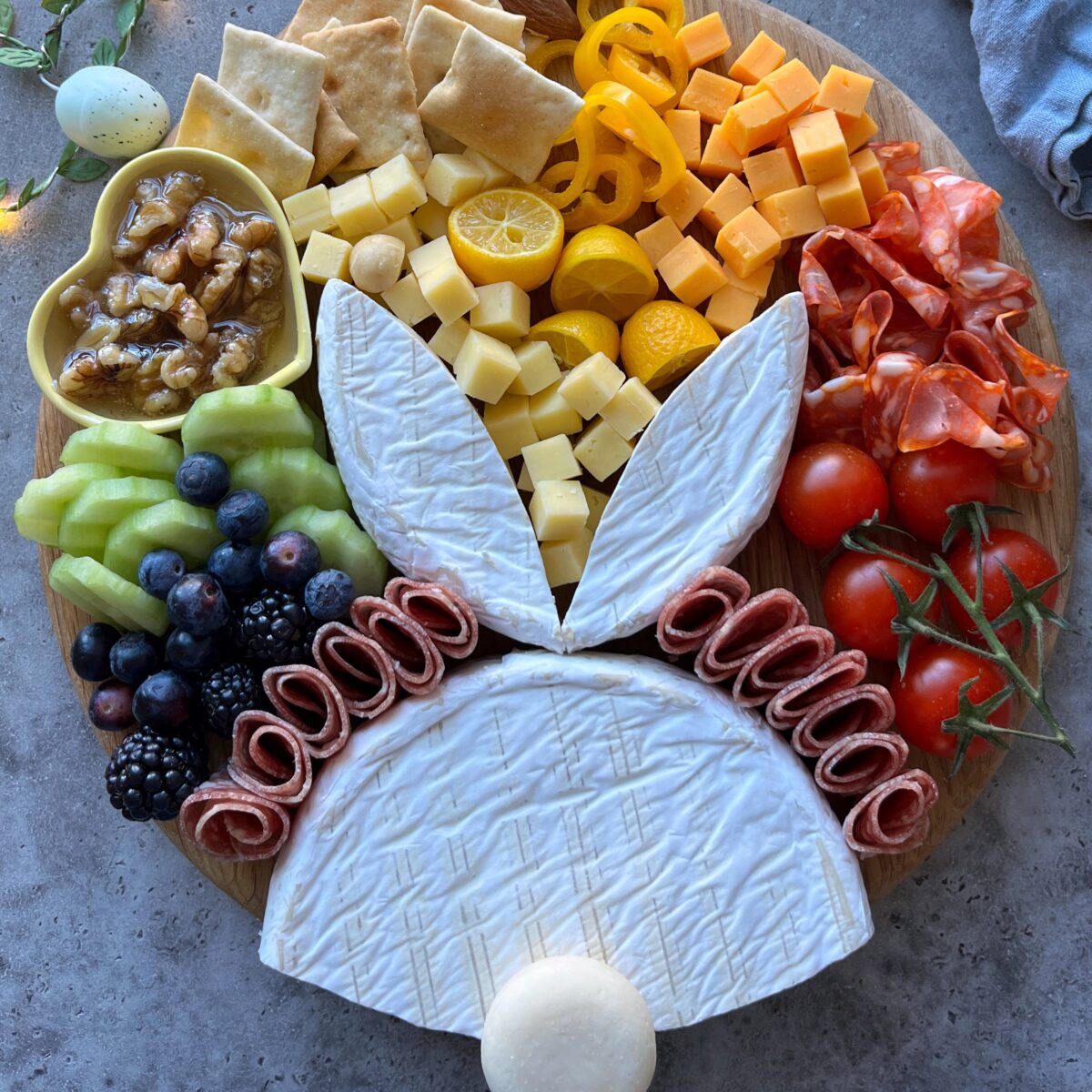 A round wooden easter charcuterie board features assorted cheeses, crackers, fruits, veggies, nuts, and meats arranged to resemble a bunny face with ears and a round white nose.