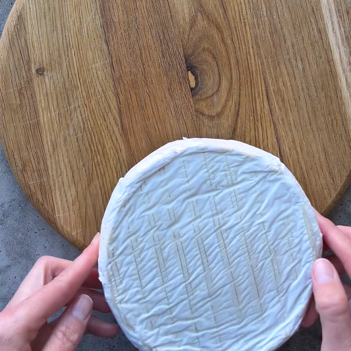 Two hands hold a wheel of soft-ripened cheese with a white rind over a round wooden cutting board.