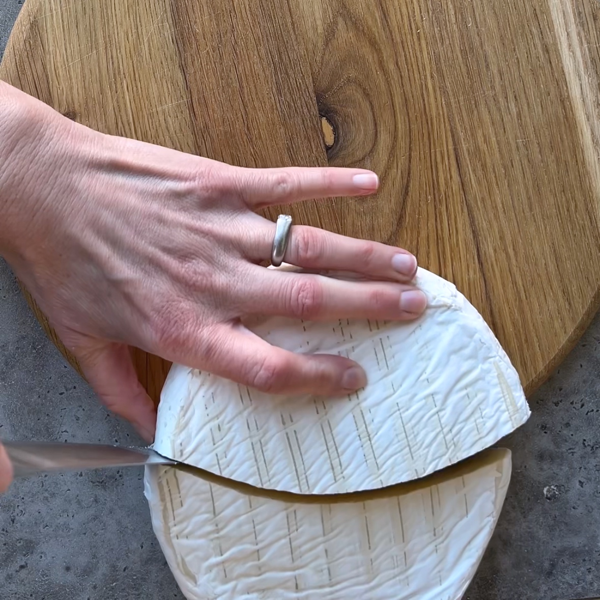 A hand holding a round of brie cheese while slicing it in half along the edge of a wooden cutting board.