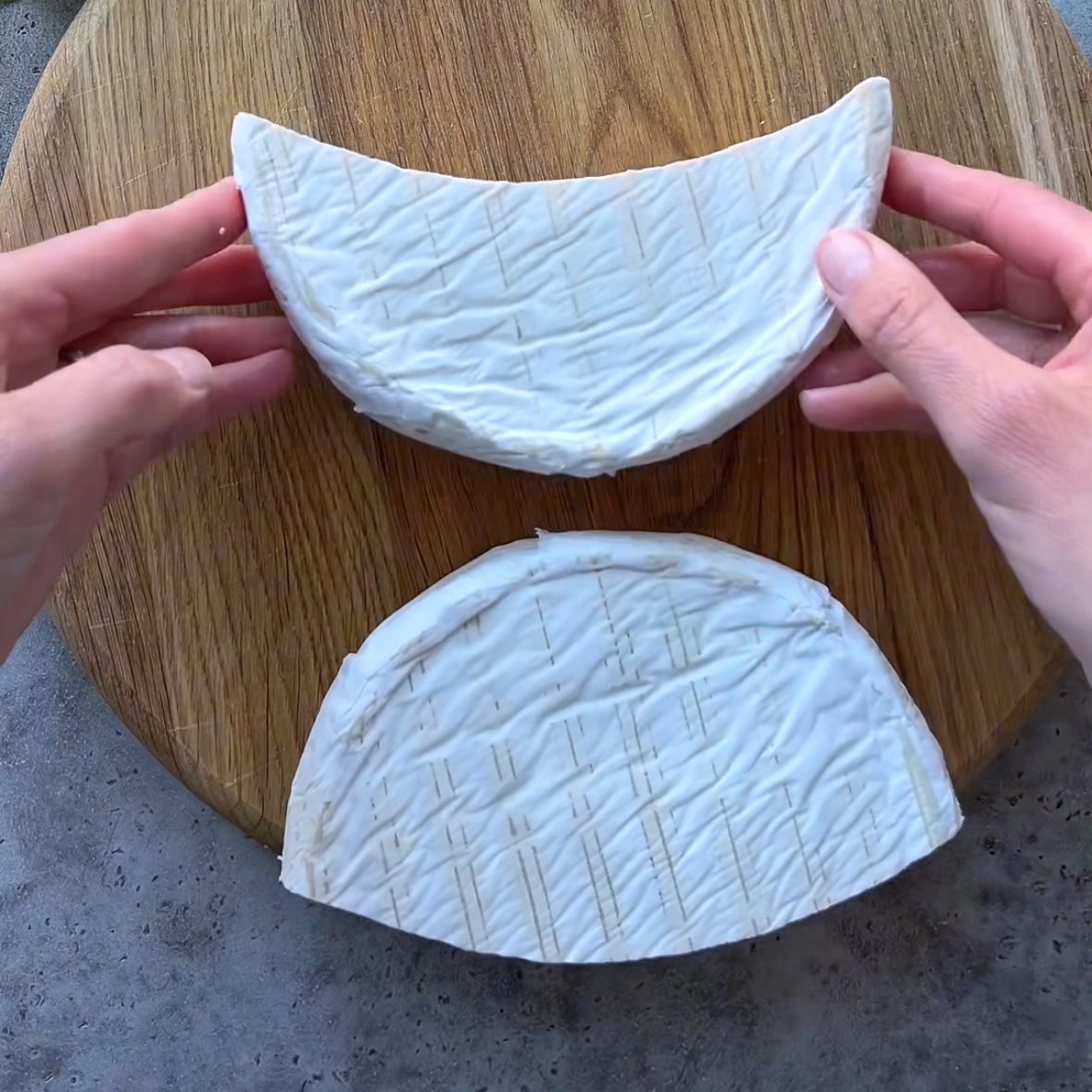 Two hands hold a wheel of brie cheese cut in half on a round wooden board, with the white rind visible.