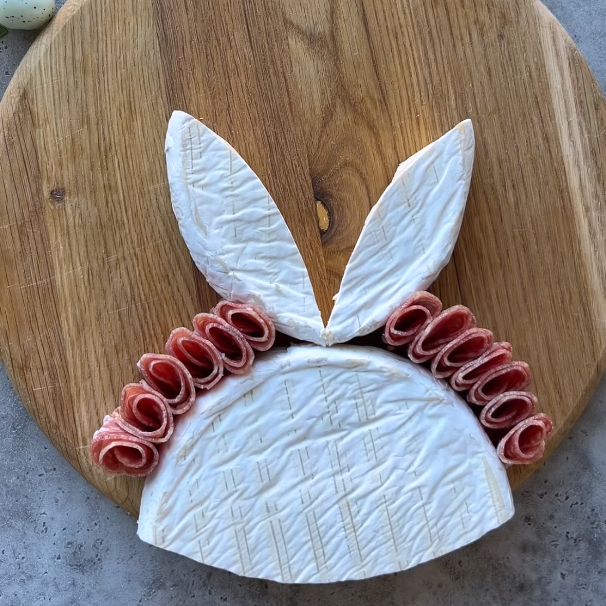 A wheel of brie cheese cut and arranged as a bunny with two ears and cured meat slices forming the sides on a wooden board.