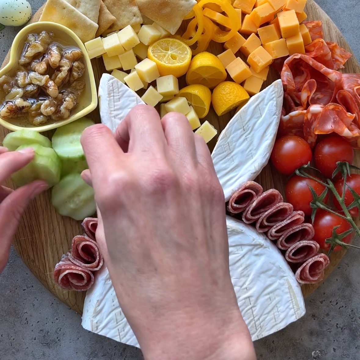 A person arranges cucumber slices on an Easter Charcuterie Board with cheese, meats, tomatoes, walnuts, crackers, and a brie shaped like a rabbit.
