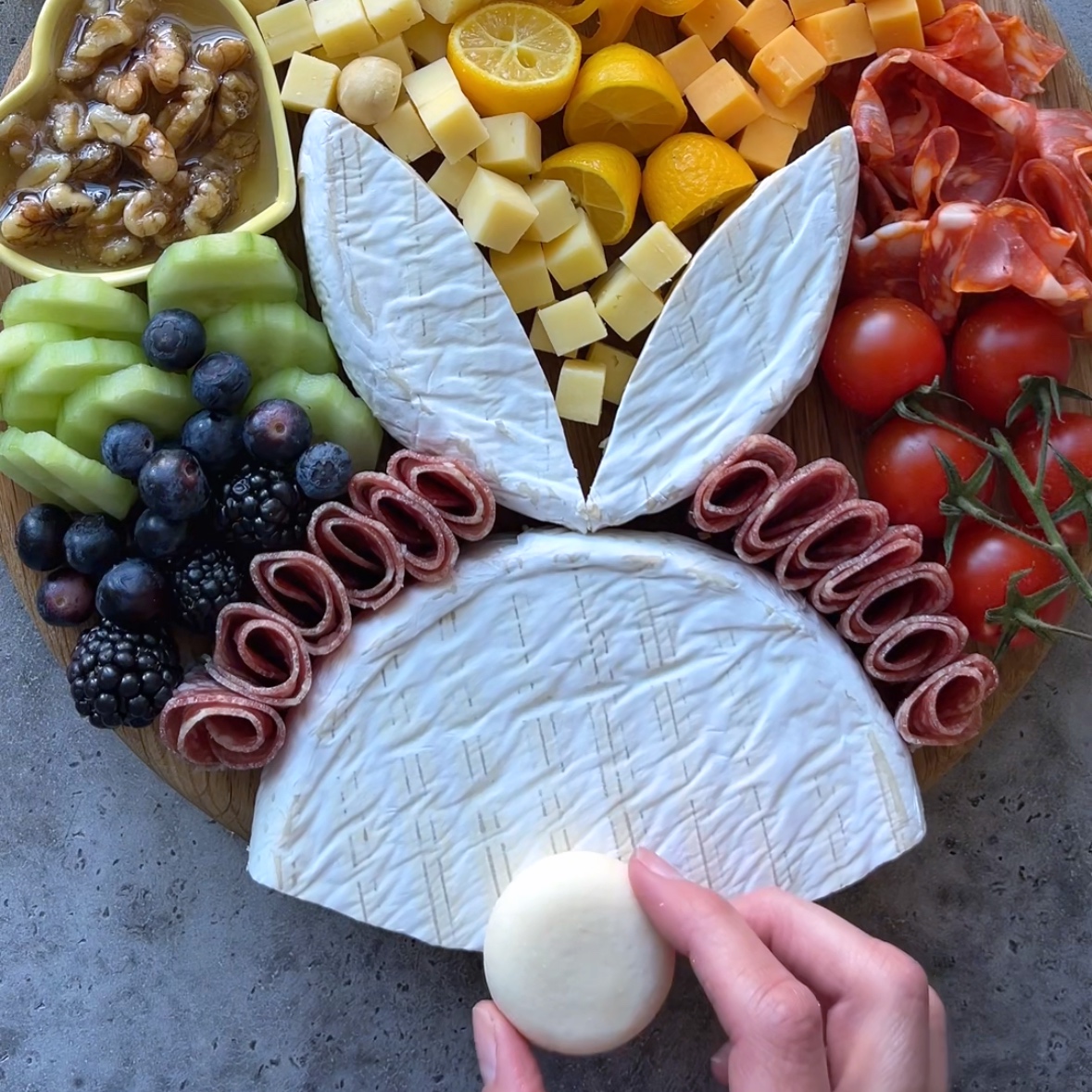 A delightful Easter Charcuterie Board is arranged in the shape of a bunny, featuring an array of assorted meats, fruits, and nuts. A hand holds a small cheese piece to create the bunnyโs tail, making this festive board perfect for your holiday gatherings.