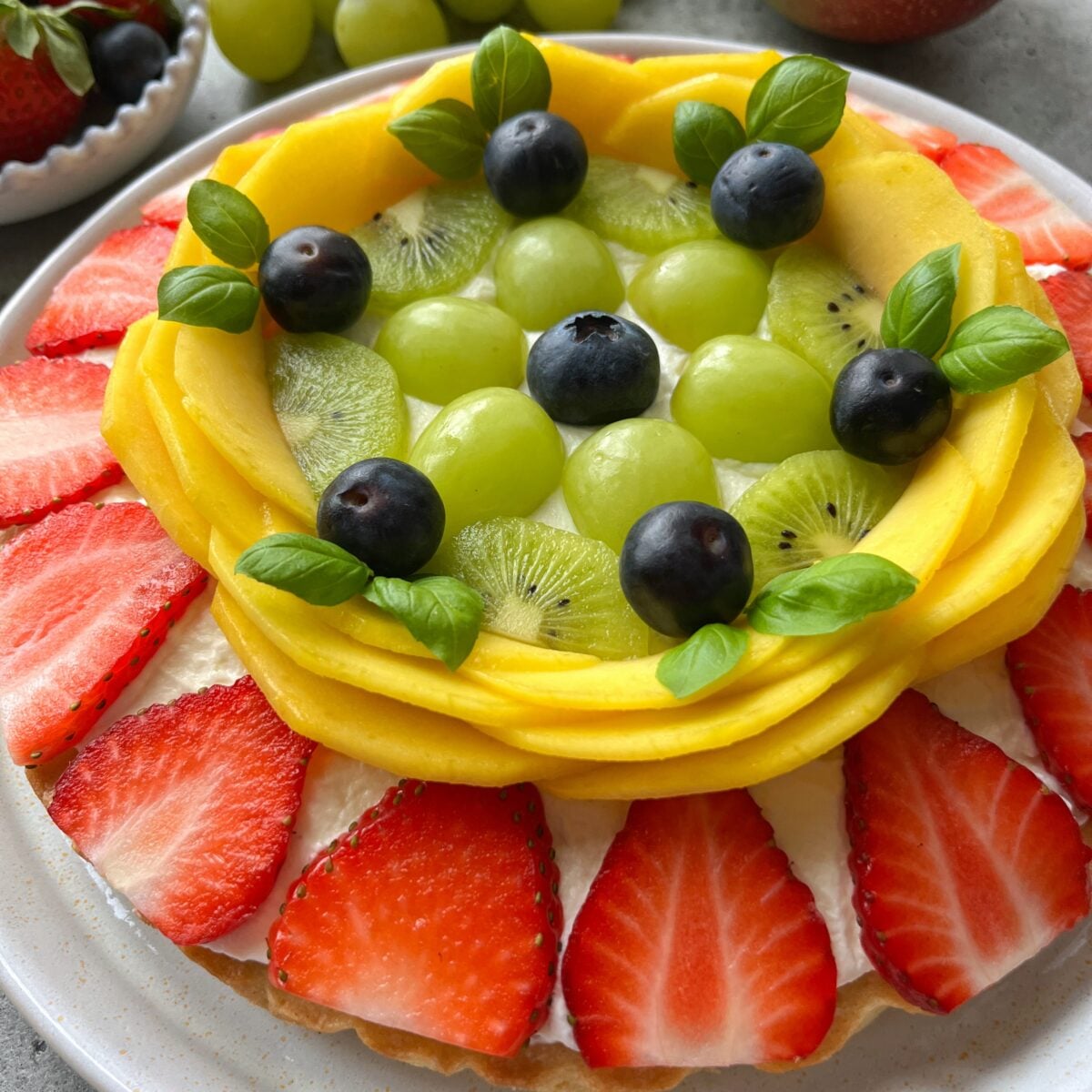 A fruit pizza topped with sliced strawberries, mango, kiwi, green grapes, blueberries, and small green leaves.
