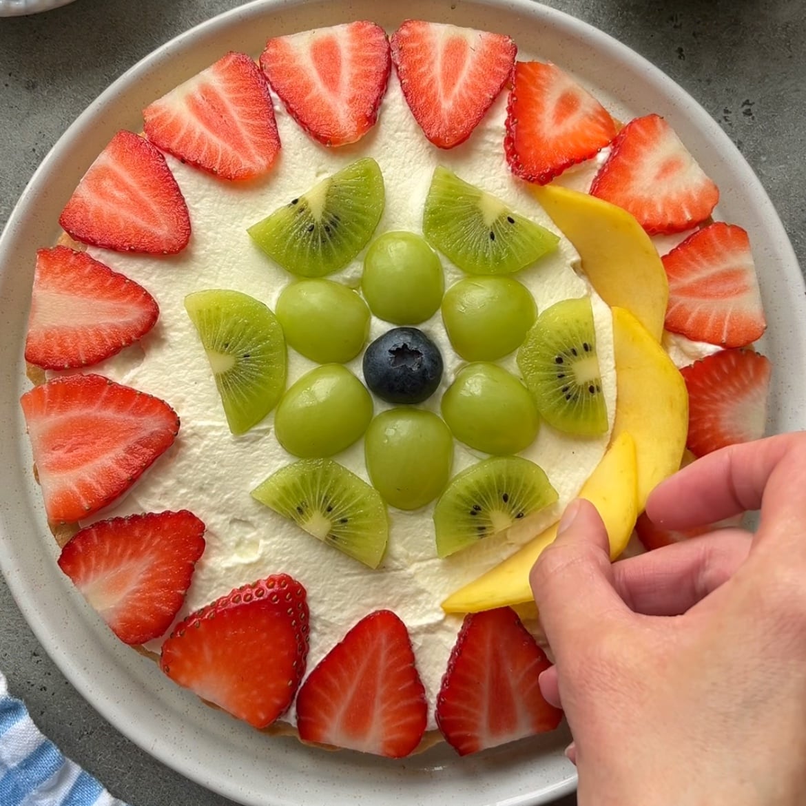 A hand places a slice of mango on a vibrant fruit pizza topped with whipped cream, strawberries, green grapes, kiwi slices, and a blueberry.
