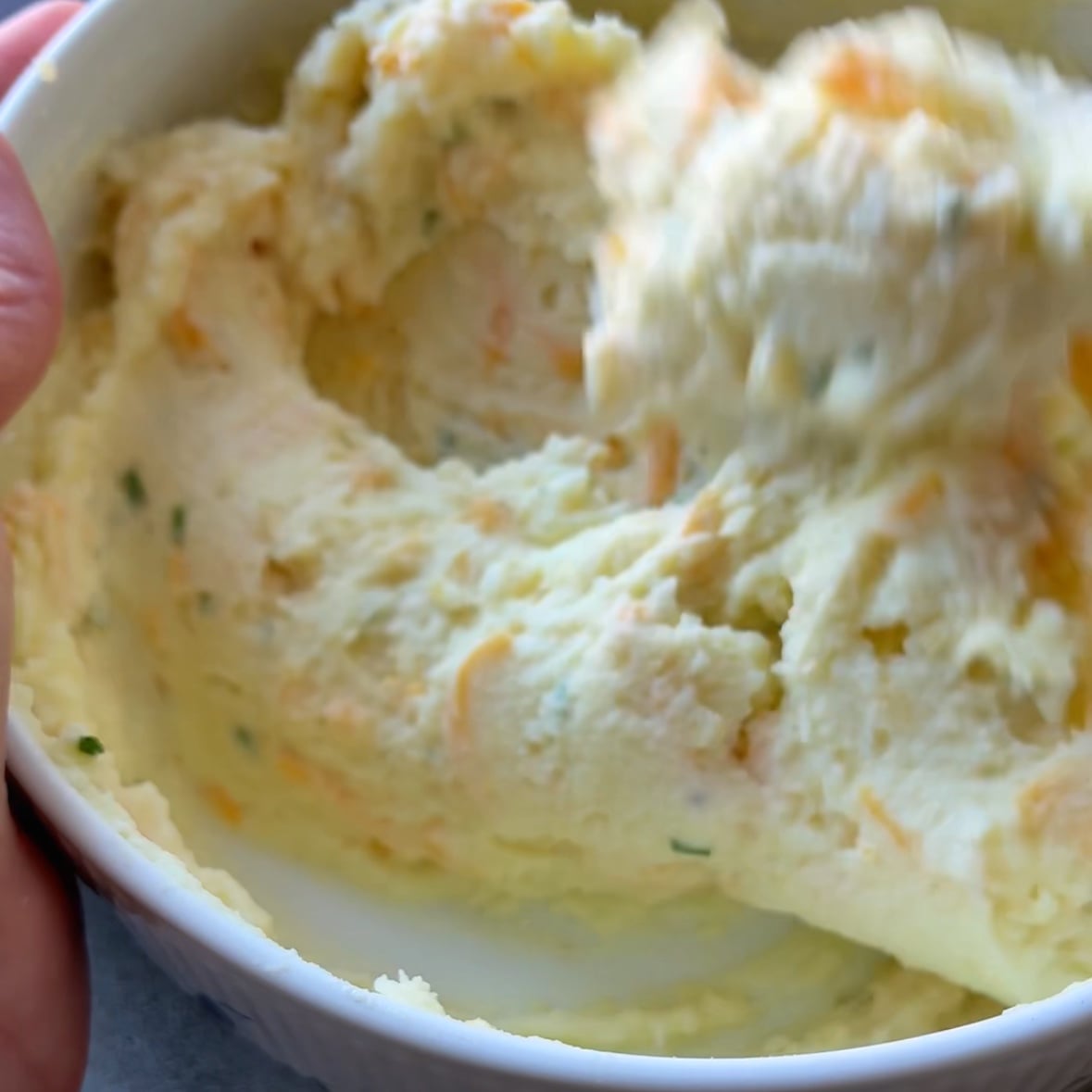 A close-up of a bowl filled with creamy mashed potatoes, showing visible bits of herbs and grated cheese being mixed in.