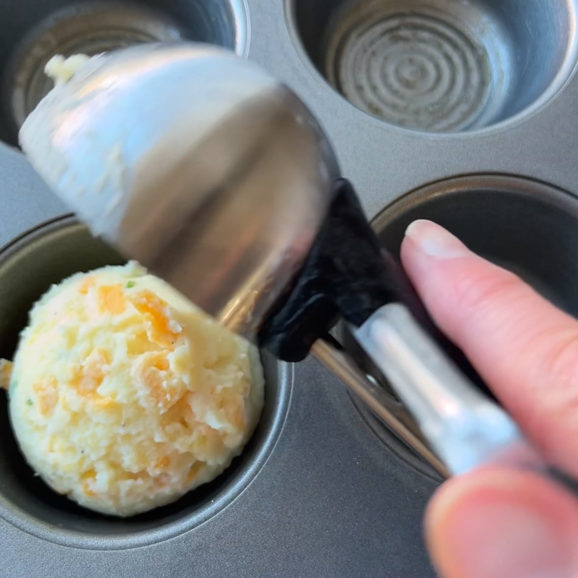A hand uses an cookie scoop to place mashed potato cup mixture into a muffin tin cavity.
