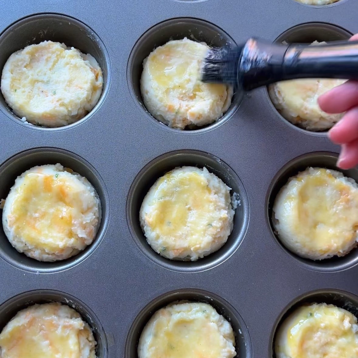 A hand uses a brush to apply an melted butter to uncooked mashed potato cups in a muffin tin.