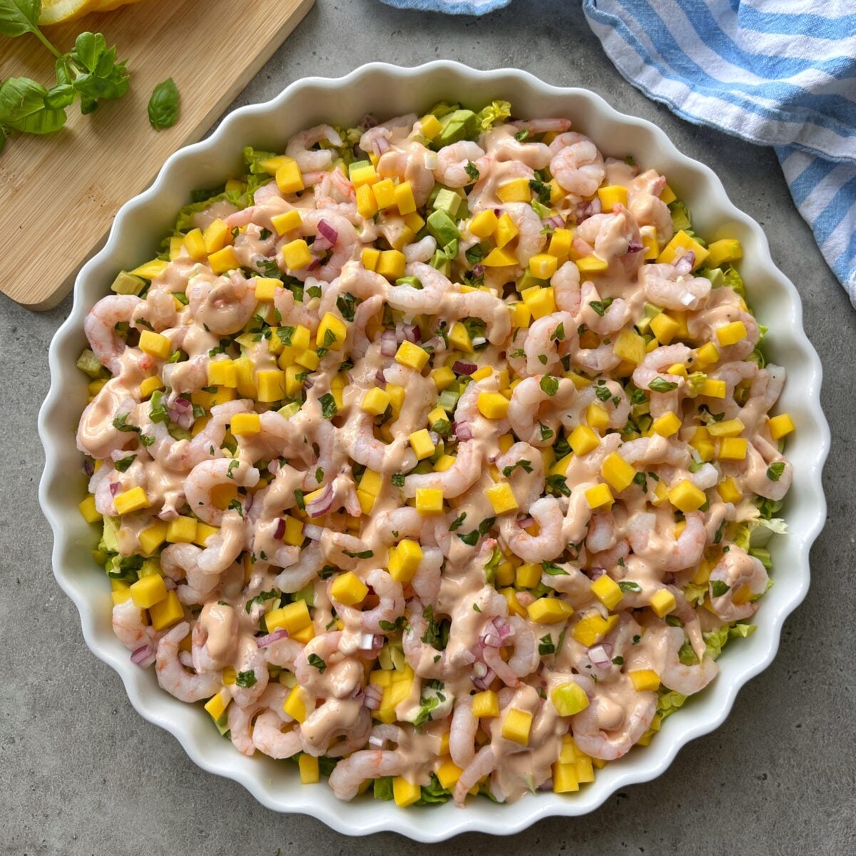 Round dish filled with Shrimp Cocktail Salad featuring shrimp, diced mango, red onion, avocado, and chopped herbs, all topped with a creamy pink dressing, on a gray surface.