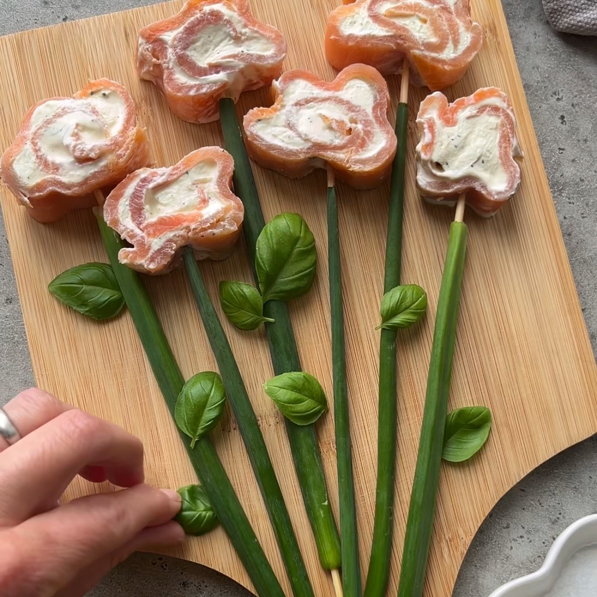 A hand holds a rolled appetizer with cream filling above a cutting board topped with green onion stalks and similar smoked salmon roulade appetizers.