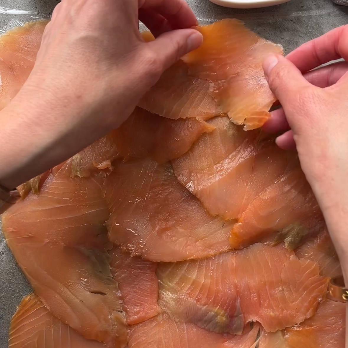 Hands arranging overlapping slices of smoked salmon on a cling film surface.
