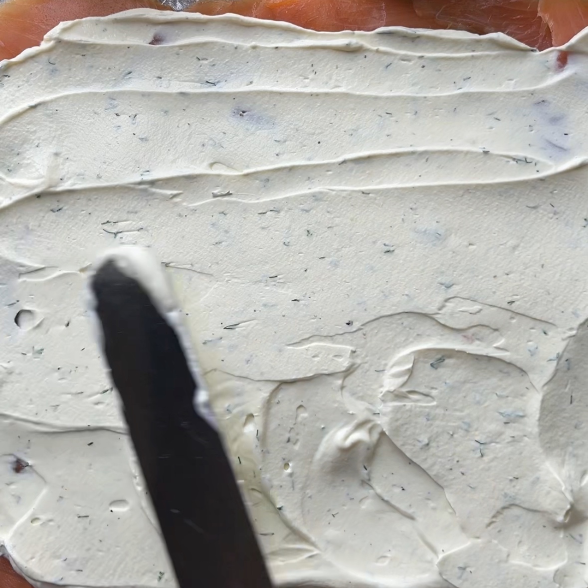 A knife spreads a layer of herb cream cheese over a layer of smoked salmon, creating smooth, textured lines.