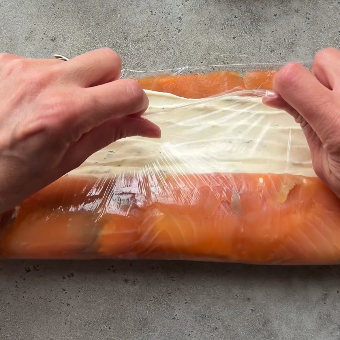 Hands roll plastic wrap around a Smoked Salmon Roulade.
