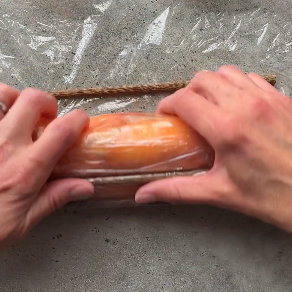 Hands wrapping a piece of smoked salmon in plastic wrap lined with chopsticks.