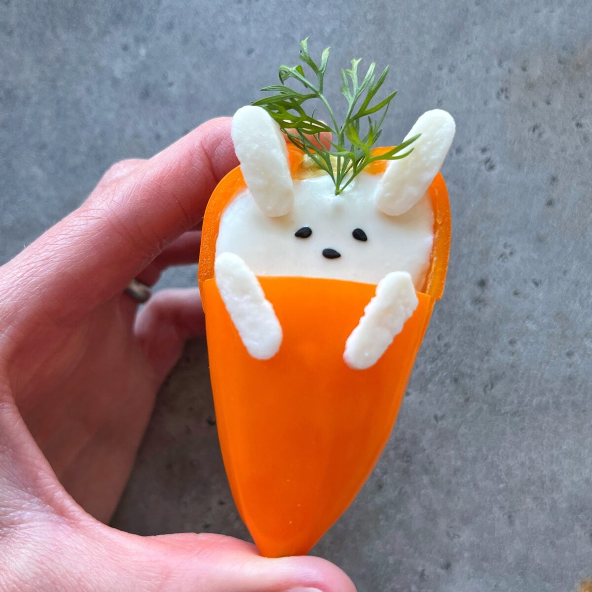 A hand holds a snack resembling a bunny made from stuffed mini peppers, with mozzarella for ears and small sesame seeds for eyes and nose, set against a grey background.