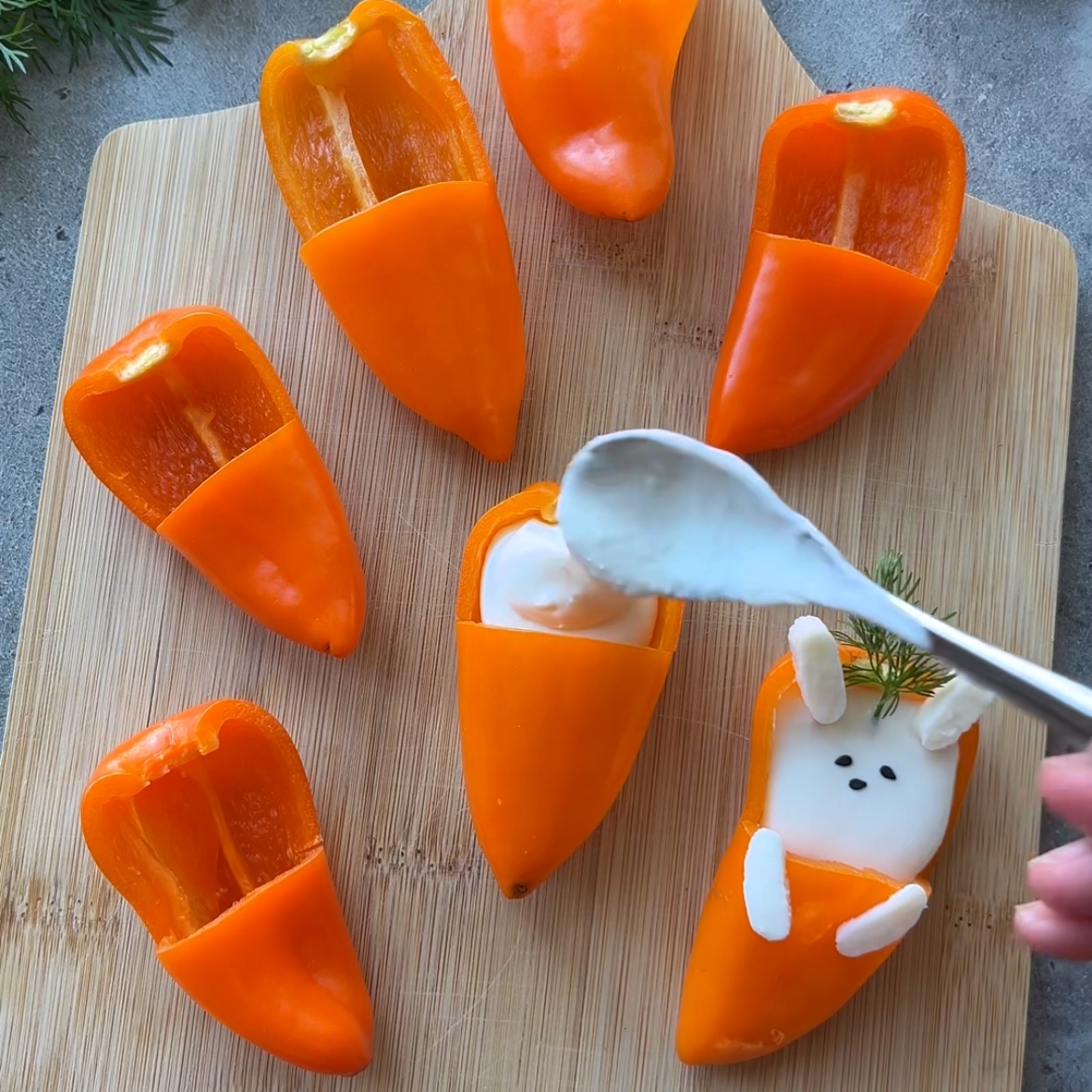 Stuffed mini peppers cut and arranged on a wooden board, some filled with whipped feta dip and decorated with herbs and shaped cheese to resemble bunnies. A spoon is adding dip to one of the peppers.