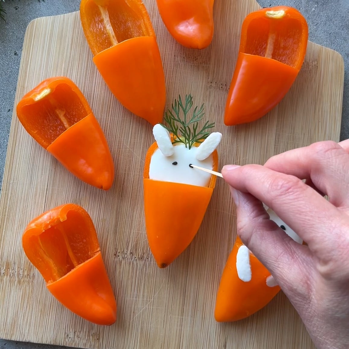 Hand decorating orange bell pepper halves filled with white filling to resemble bunnies, using herbs for ears, on a wooden cutting board.