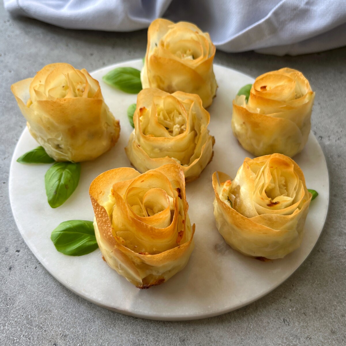 Six golden, rose-shaped phyllo appetizers are arranged on a round white plate with fresh basil leaves, set on a gray surface with a light cloth in the background.