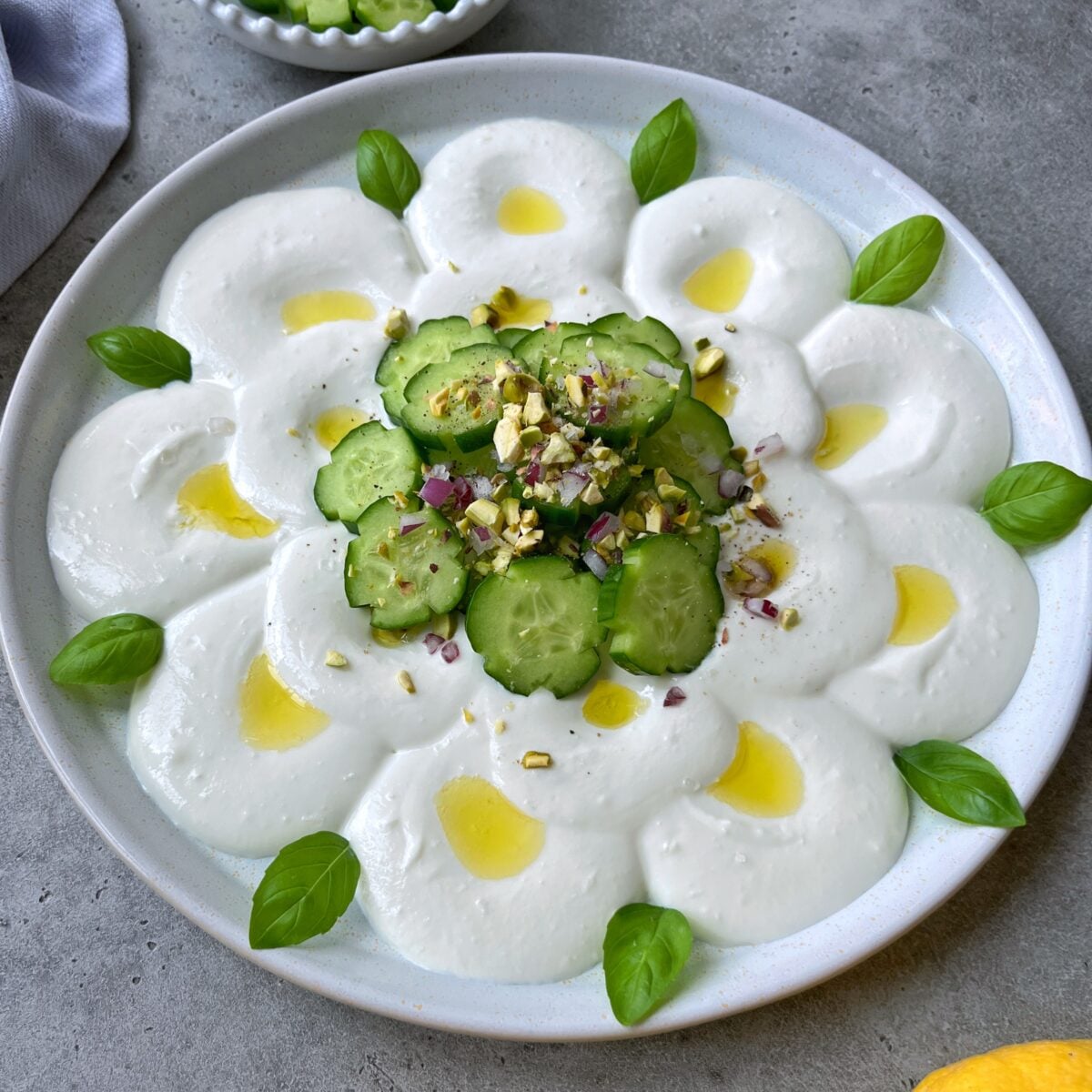 A plate of creamy Feta Cheese Dip decorated with sliced cucumbers, chopped nuts, diced red onions, olive oil, and fresh basil leaves.