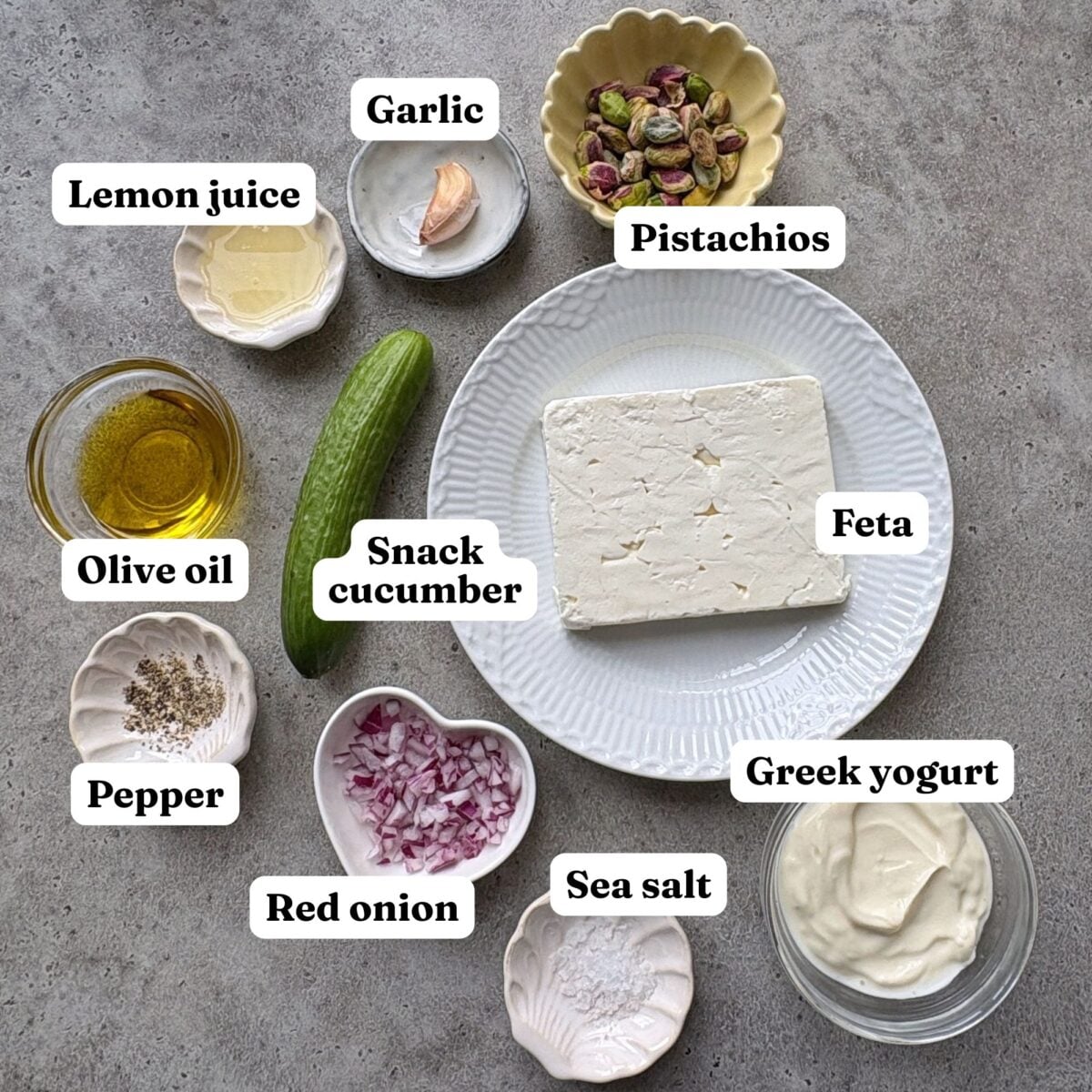 Assorted labeled ingredients for a Feta Cheese Dip, including feta, Greek yogurt, olive oil, lemon juice, garlic, pistachios, snack cucumber, red onion, pepper, and sea salt, arranged on a table.