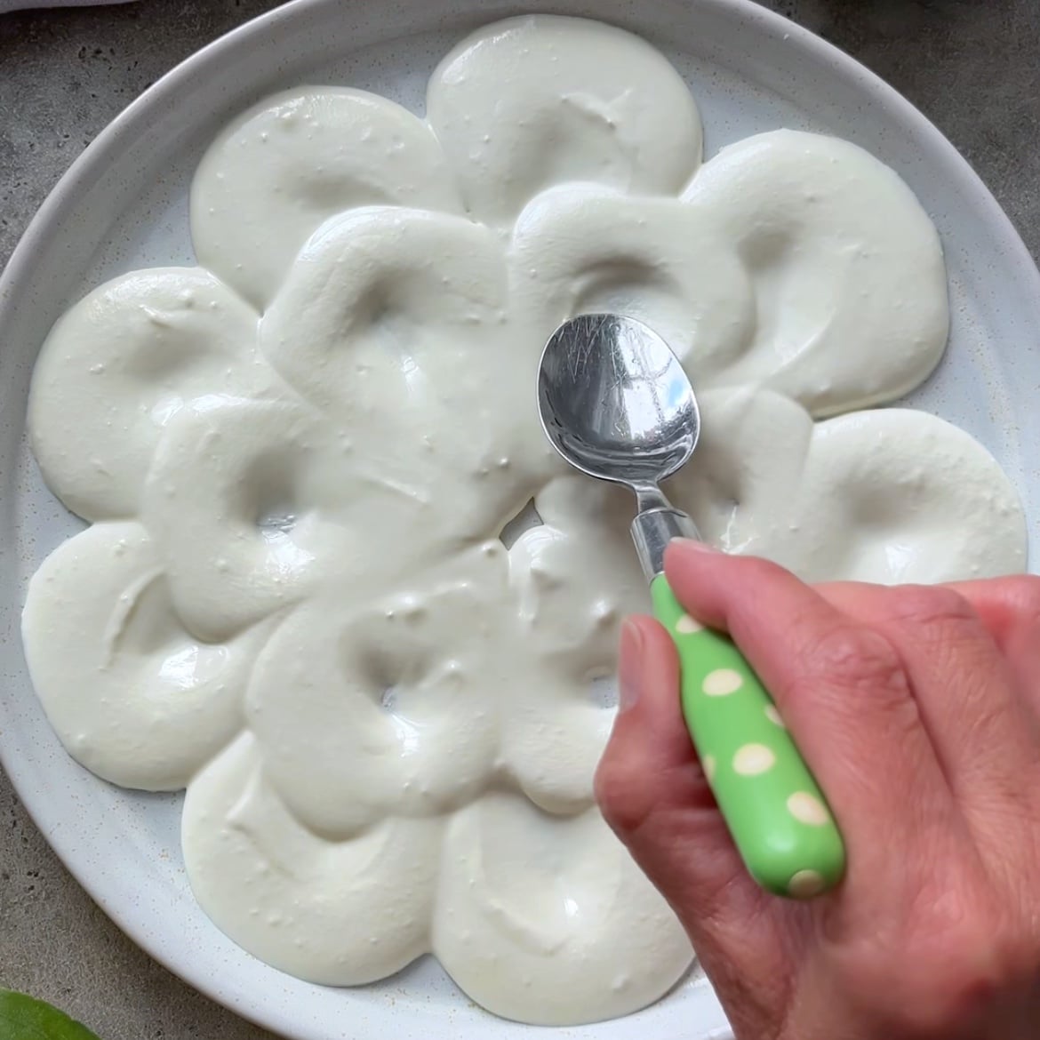 A hand uses a spoon with a green polka-dot handle to spread creamy Feta Cheese Dip in petal shapes on a plate.