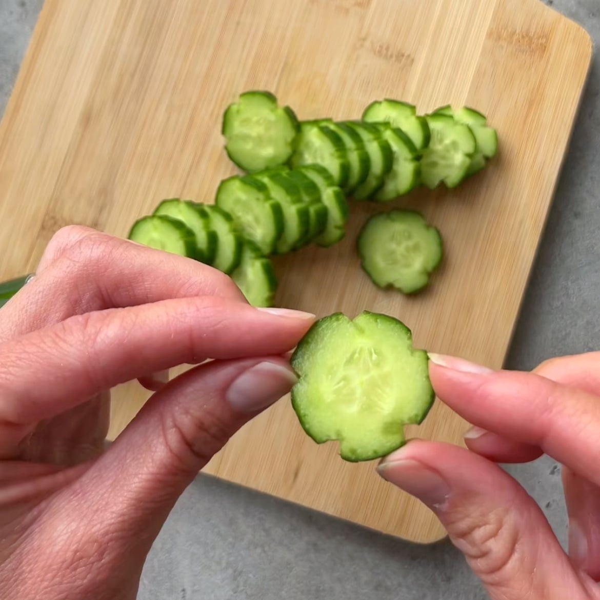 Hands holding a scalloped cucumber slice in front of a cutting board with more scalloped cucumber slices.